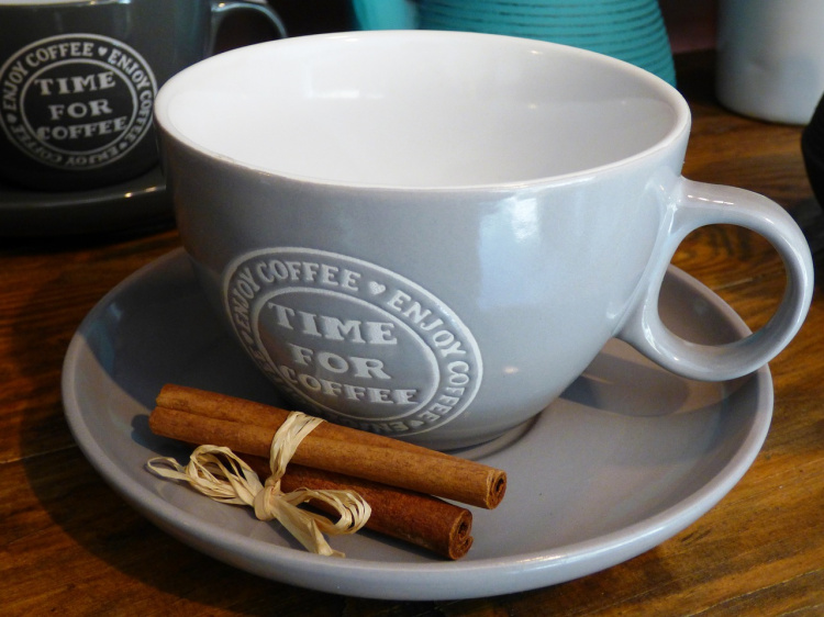 Kuvassa harmaa kahvikuppi jossa lukee time for coffee kupin edessä on kolme kanelitankoa