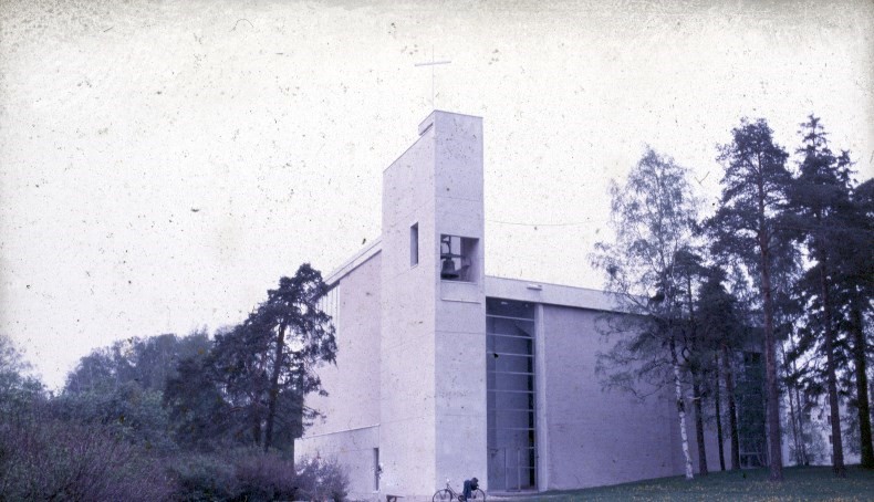 Kirkon takapuolelta otettu vanha valokuva. Katolla iso risti sekä kirkonkello etualalla.