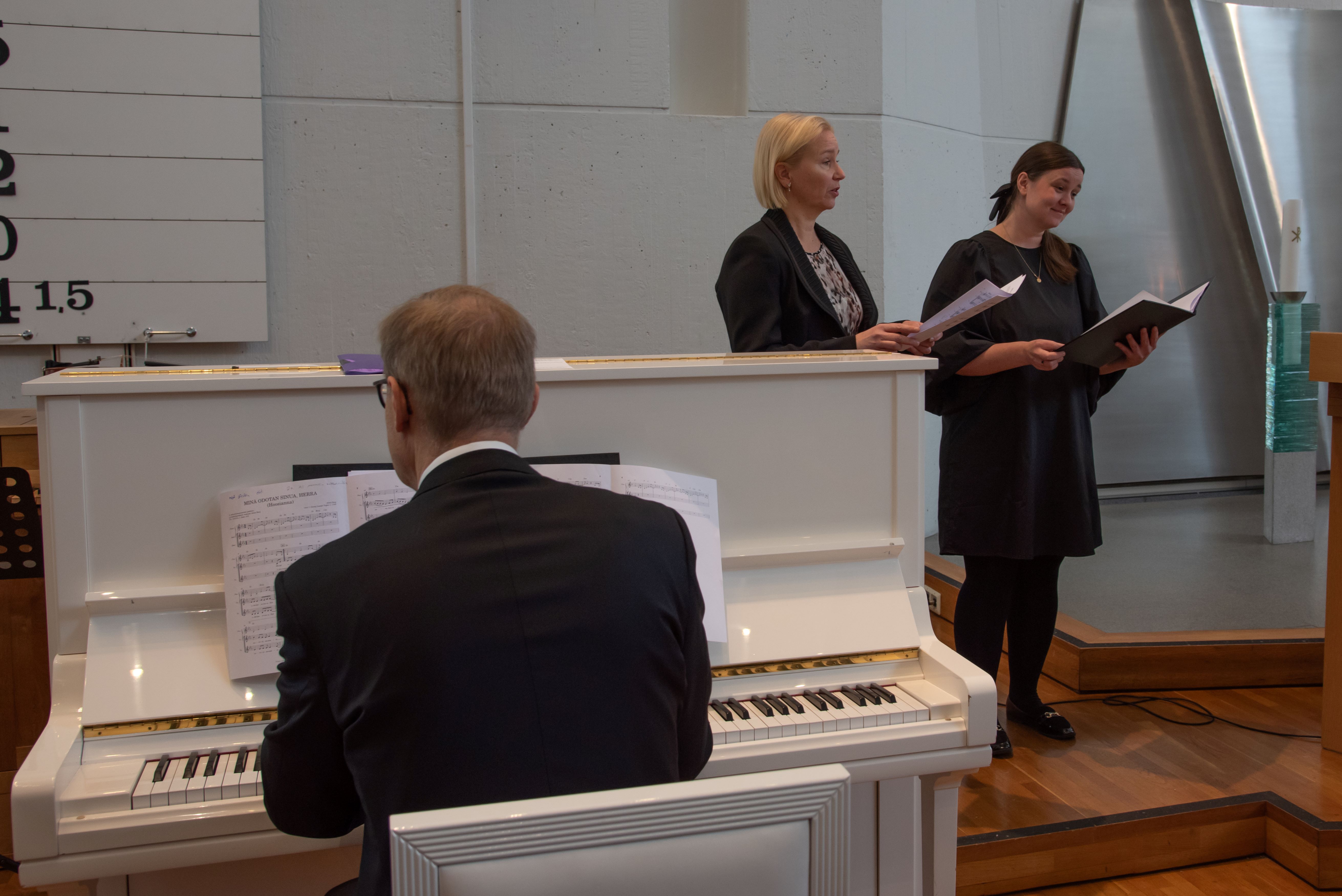 Janne Kaksonen selin pianossa ja Heli Kaasalainen ja Katariina Kopsa laulamassa.