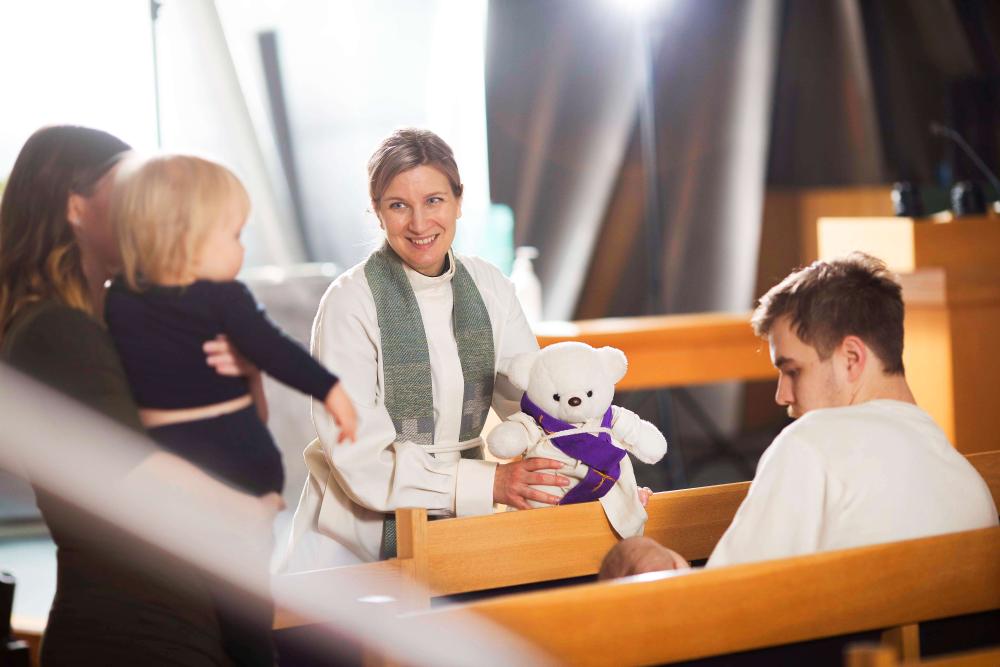 Kuvassa hymyilevä pappi ja lapsi vanhempineen ja linkki vie kirkon rekrytointijärjestelmään.