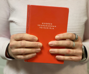 punainen laulukirja henkilön kädessä, laulukirjan kannessa teksti nuoren seurakunnan veisukirja